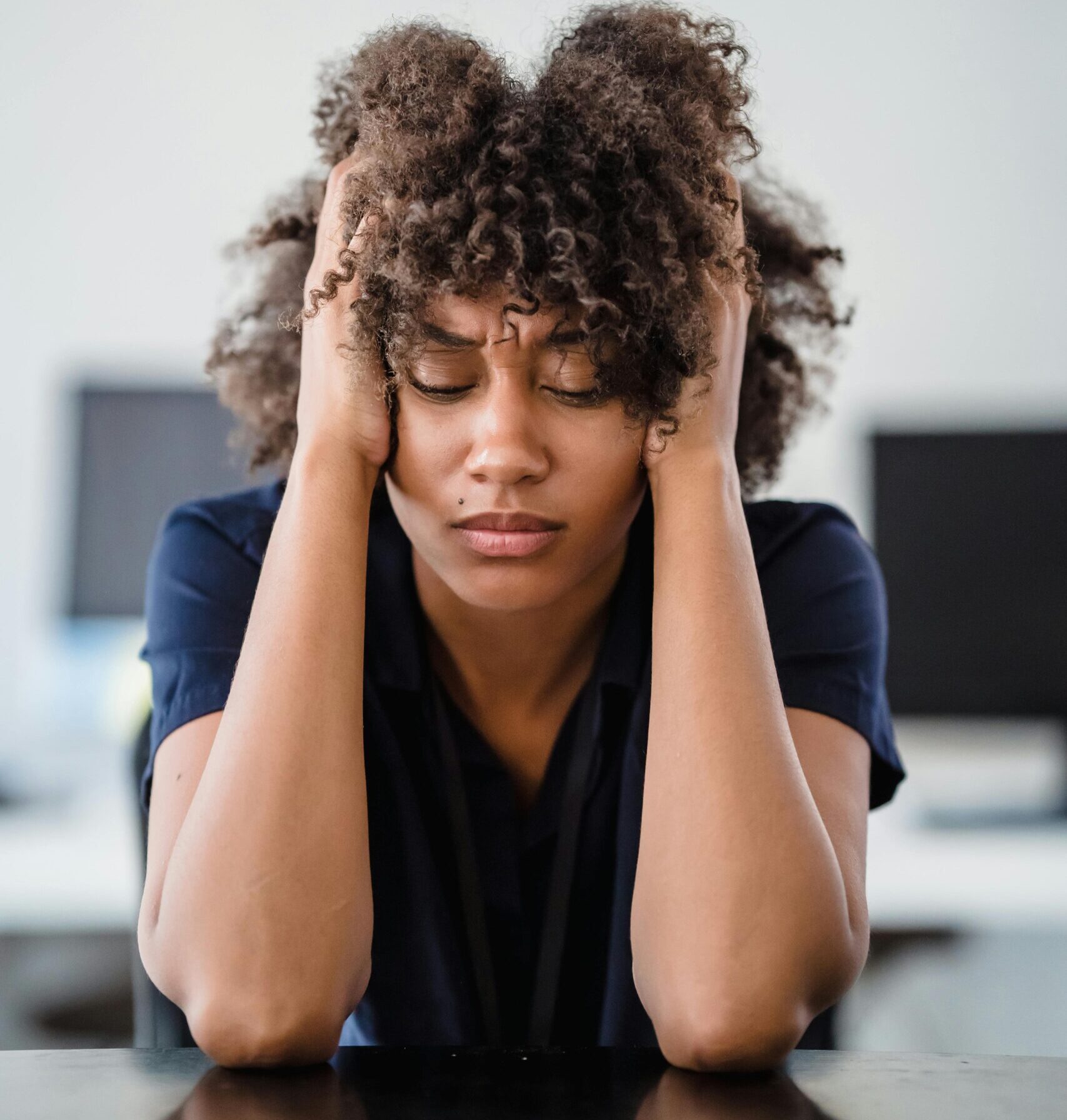 Woman in office setting expresses stress, seated at desk with hands in hair. Perfect for workplace stress concept.