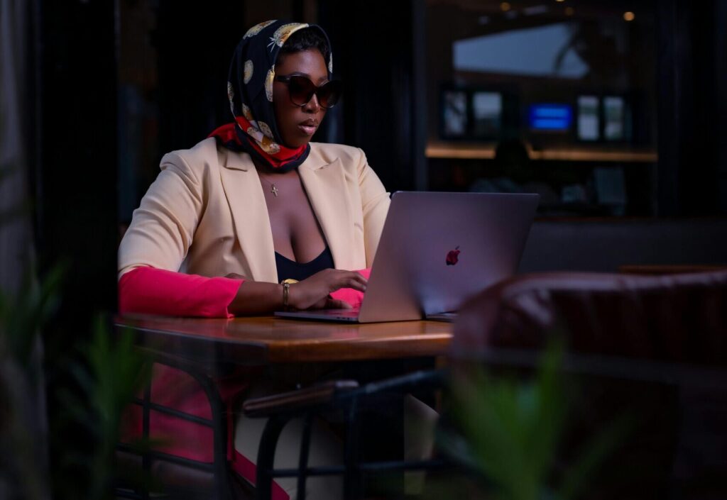 Chic woman wearing scarf and sunglasses working on a laptop in a cozy cafe setting.